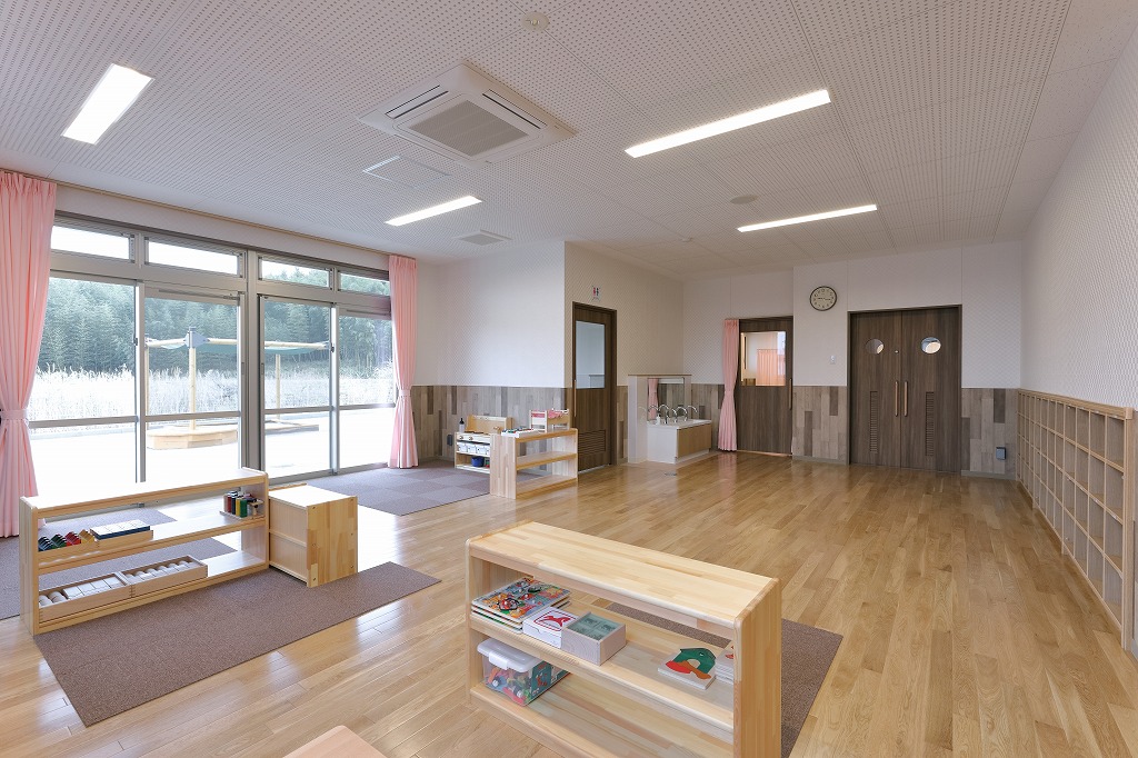 大きな掃き出し窓から園庭が見える、木目調の腰壁とピンクのカーテンが特徴的な真愛三ツ谷こども園の2歳児保育室