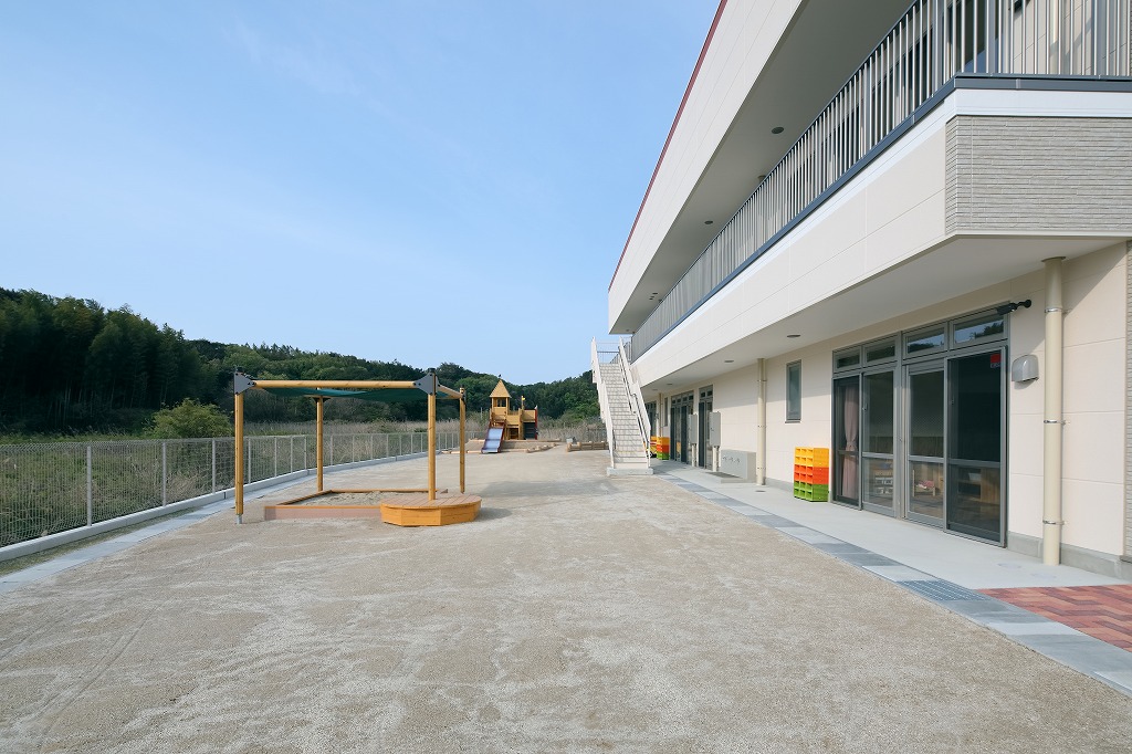 手前に日よけ付きの木製砂場を配し、奥に豊かな山の緑が広がる真愛三ツ谷こども園の広大な園庭と園舎外観。
