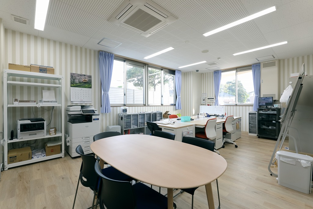 個々の事務デスクと収納棚が並び、窓からの自然光が差し込む真愛三ツ谷こども園の広々とした職員室事務エリア。